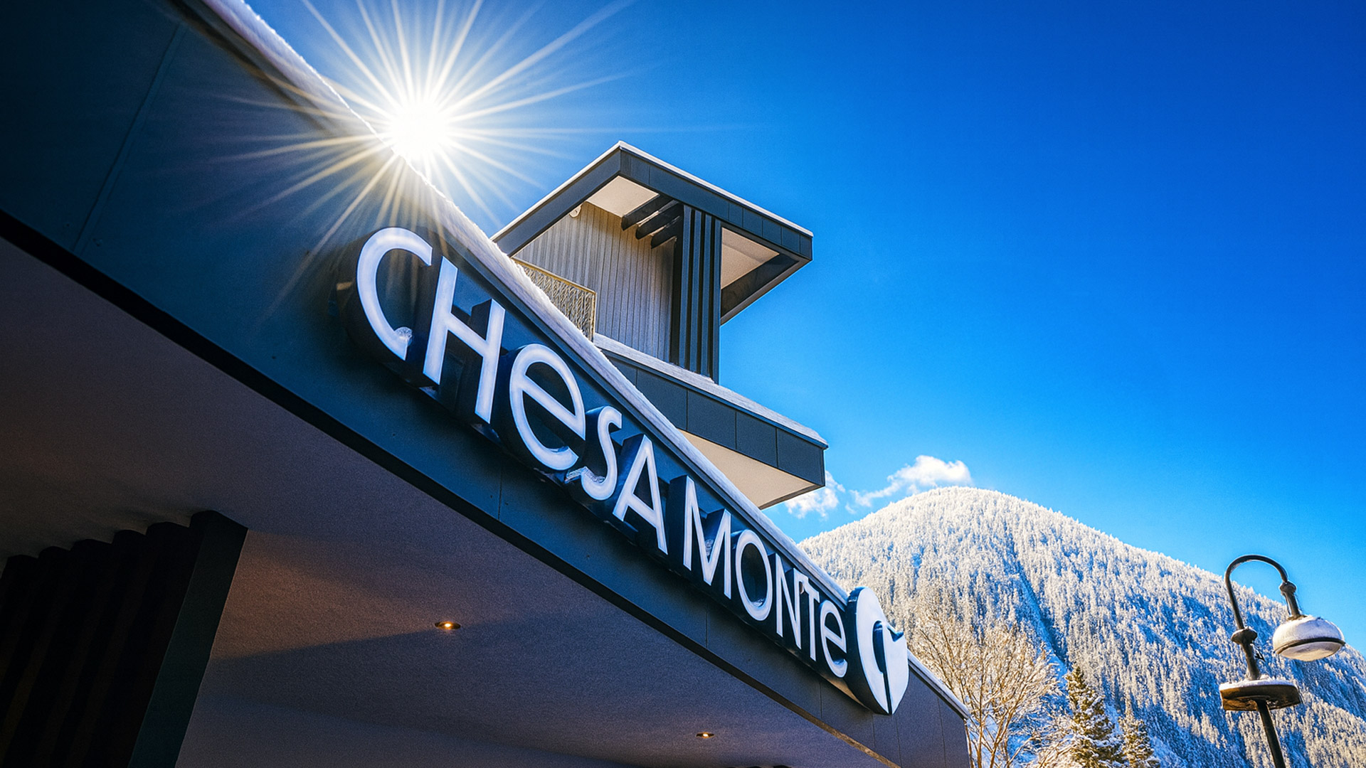Nahaufnahme des Hotel-Schriftzugs „CHESA MONTE“ an der Fassade, mit Sonnenstern im oberen Bereich und schneebedeckten Bergen vor tiefblauem Himmel im Hintergrund.