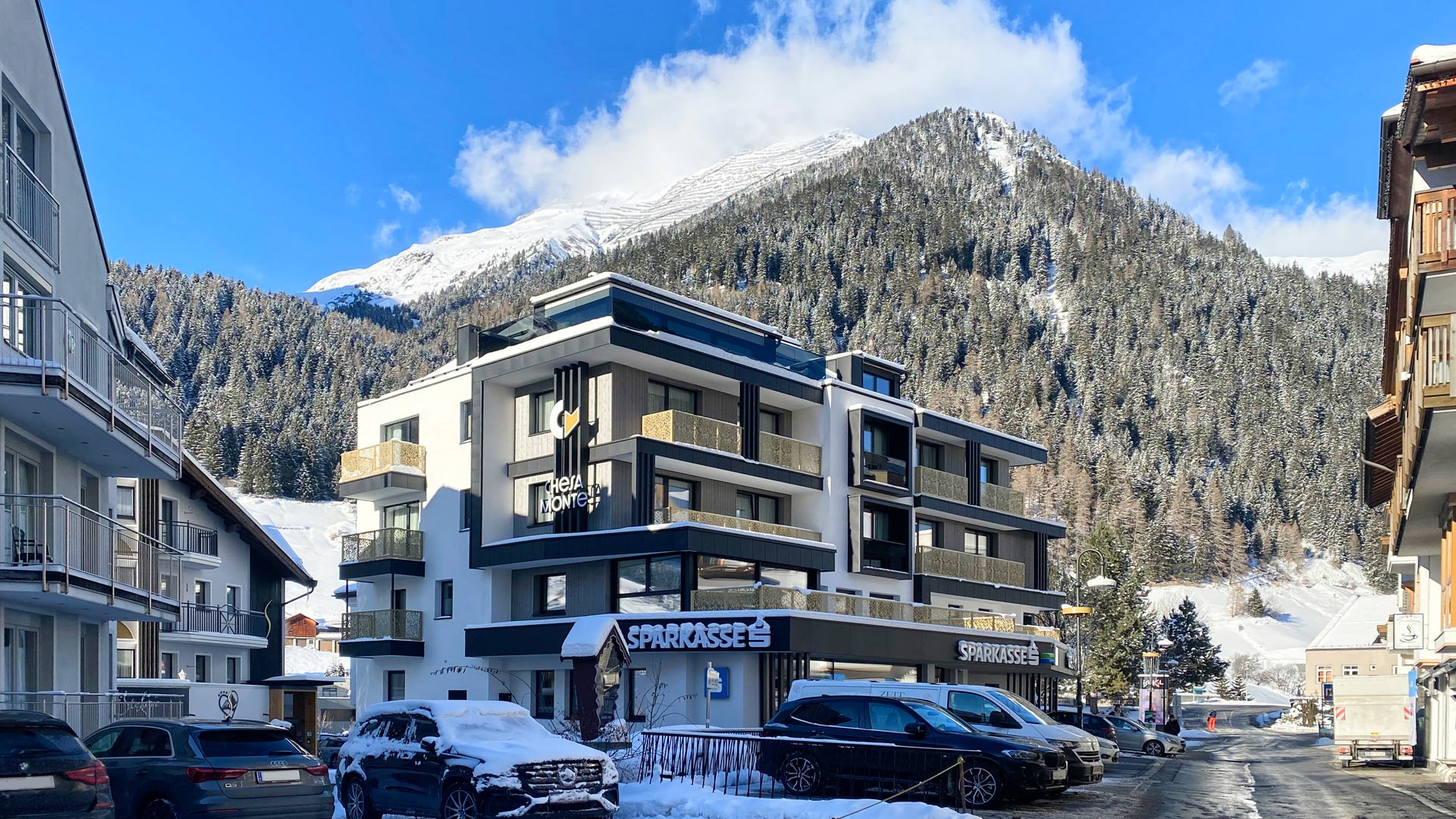 Winterliche Ortsstraße mit schneebedeckten Hotels und geparkten Autos, im Hintergrund ein hoher, verschneiter Berg unter blauem Himmel.