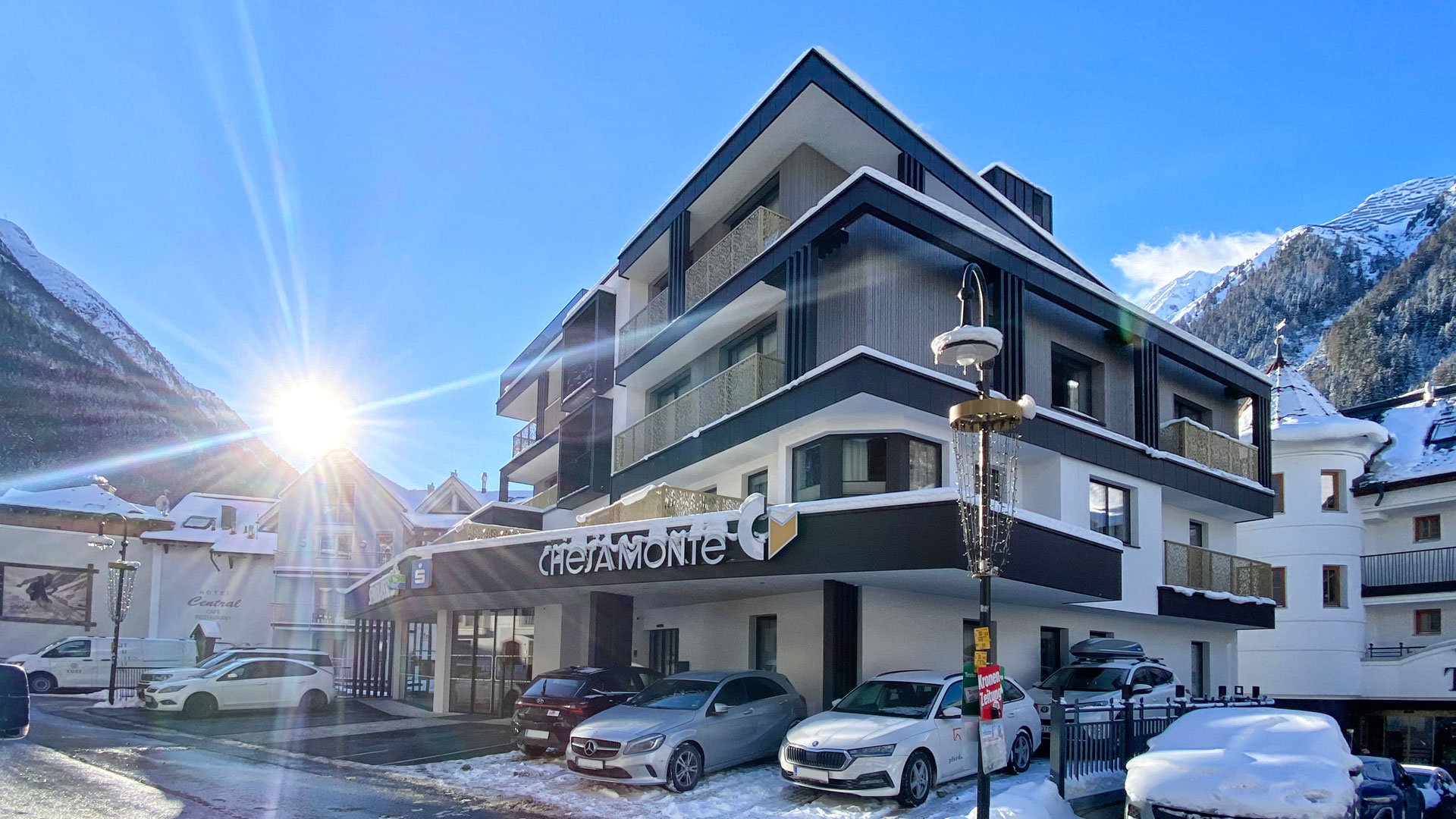 Modernes Hotelgebäude in verschneiter Winterkulisse mit geparkten Autos vor dem Eingang, strahlende Sonne links im Bild und schneebedeckte Berge im Hintergrund.