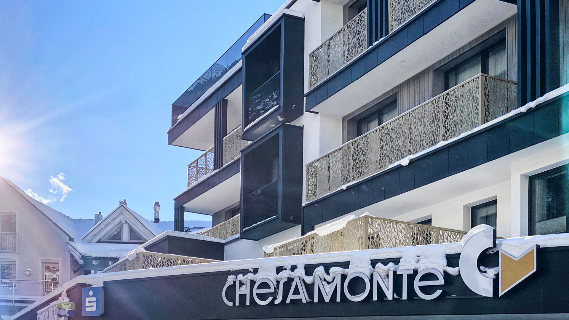 Detailansicht der modernen Hotelfassade mit dunklen Balkonen und dem Schild „CHESA MONTE“, links Sonne im Winterhimmel und schneebedeckte Berge im Hintergrund.