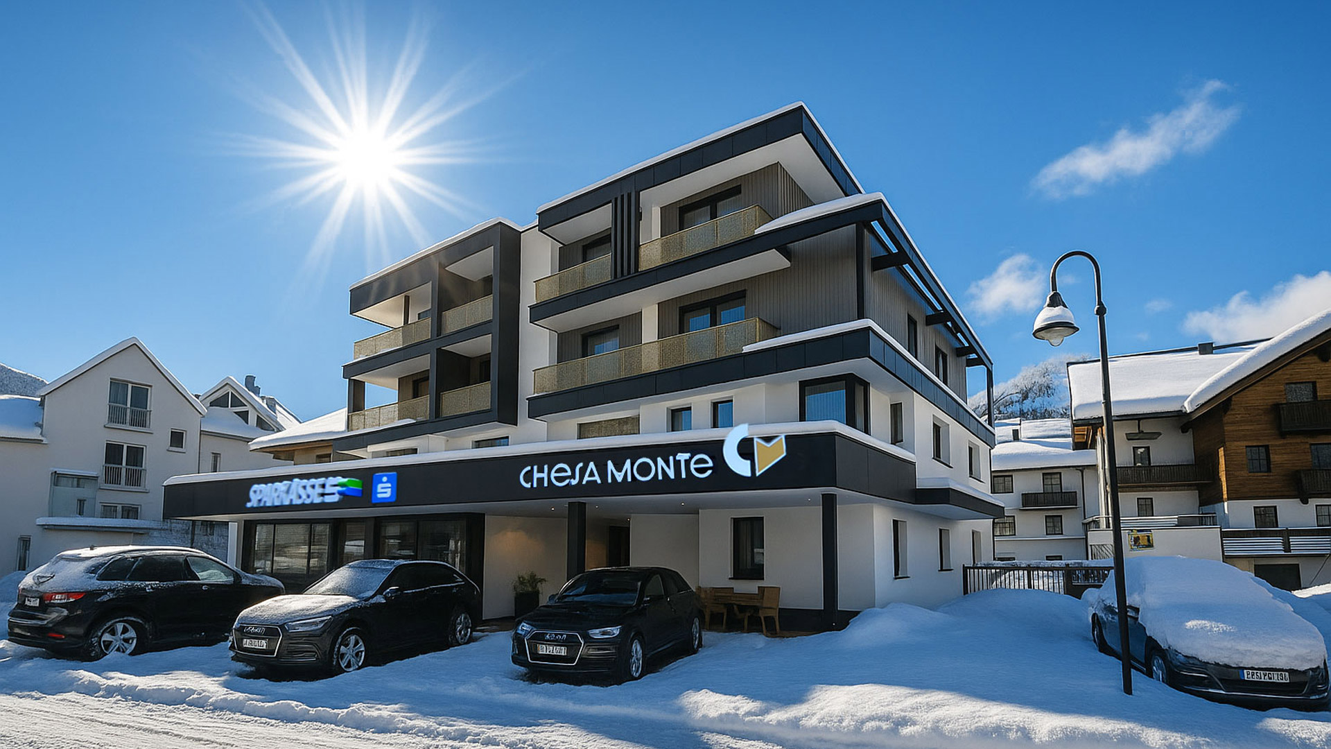 Verschneites Chesa Monte mit mehreren Balkonen und geparkten Autos vor dem Eingang, strahlender Sonnenschein am blauen Winterhimmel.