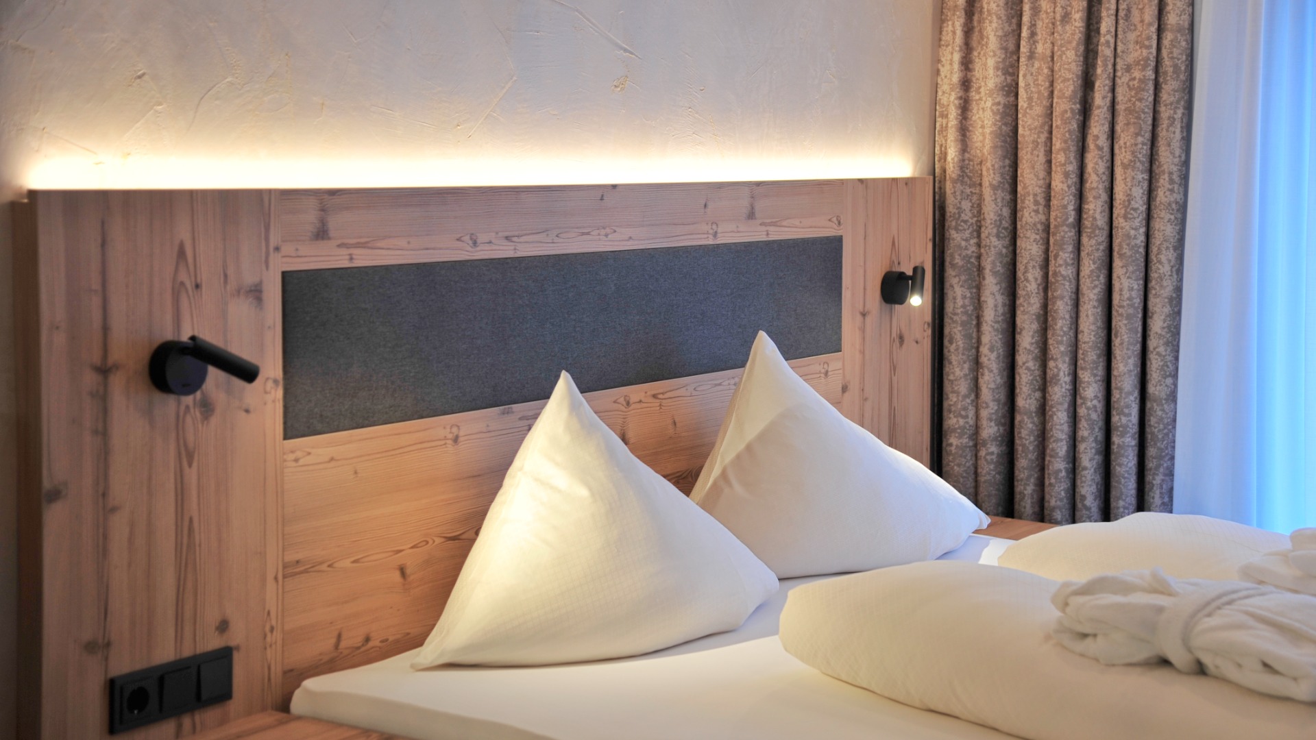 Detailaufnahme des Bettes mit Holzkopfteil, Leselampen und strukturiertem Vorhang in elegantem Studioambiente.