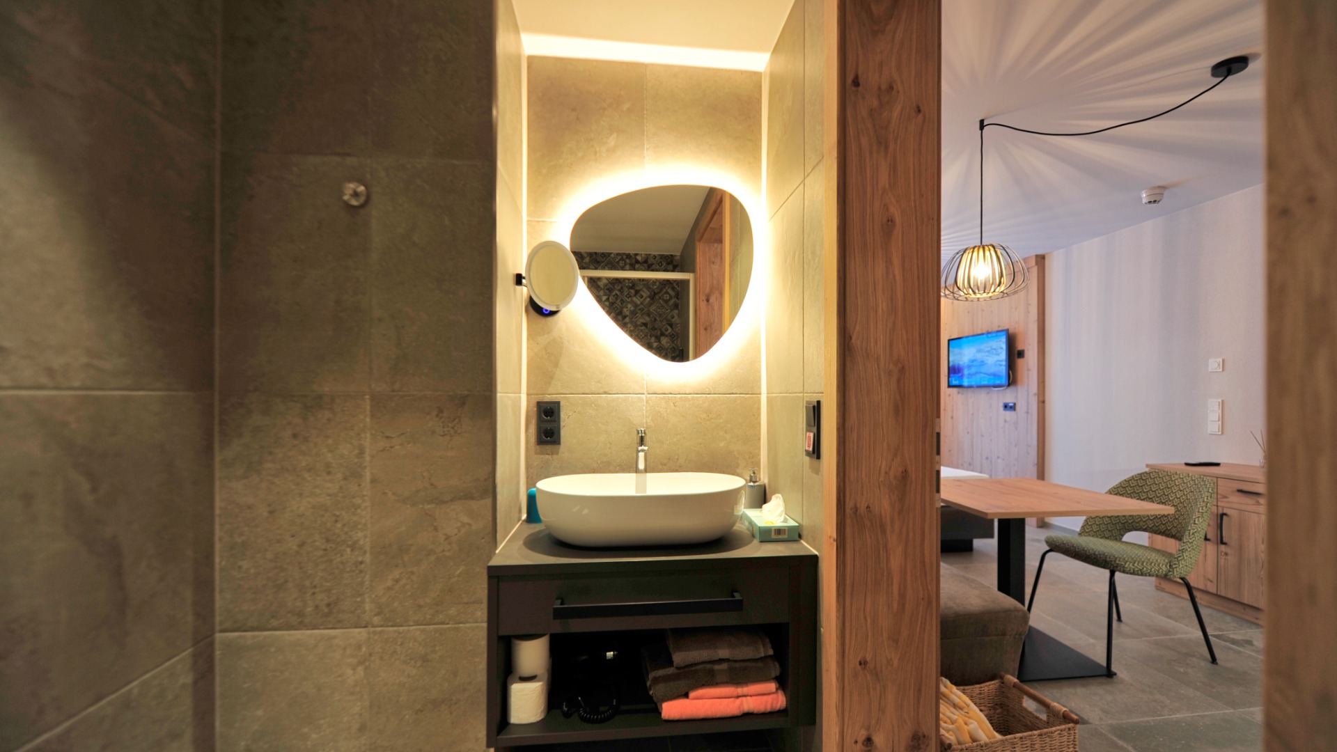 Badezimmer mit beleuchtetem Spiegel, Aufsatzwaschbecken und Blick in den modernen Wohnraum mit Esstisch.