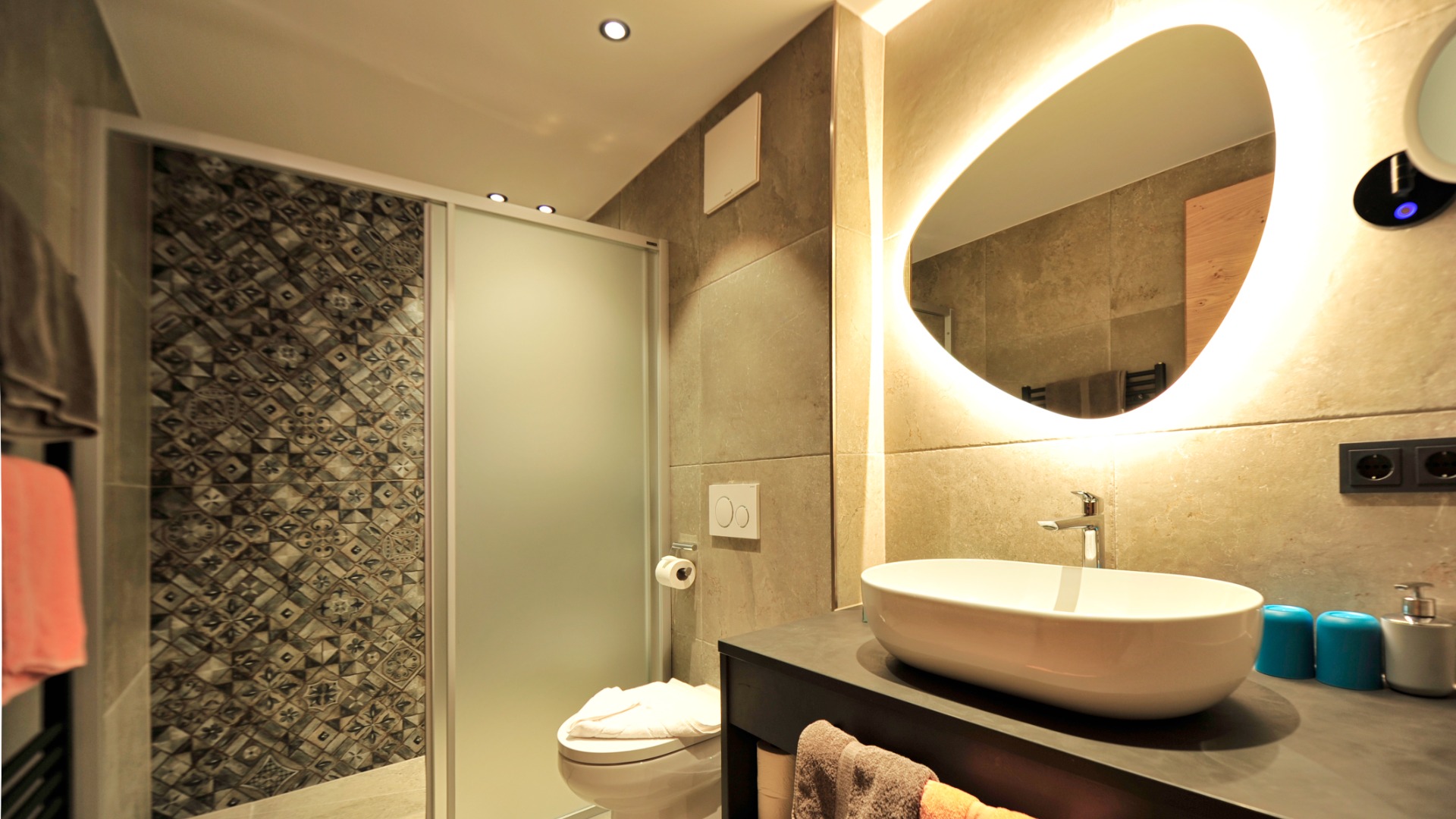 Modernes Bad mit beleuchtetem Spiegel, Aufsatzwaschbecken und Dusche mit gemusterter Fliesenwand.