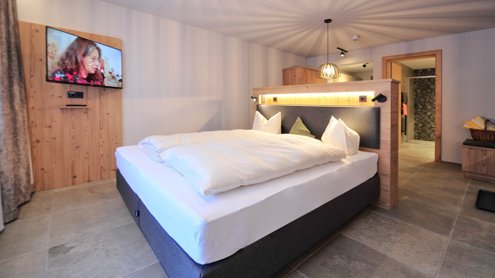 Komfortables Doppelbett mit TV an Holzverkleidung, modernem Lichtkonzept und direktem Zugang zum Bad.